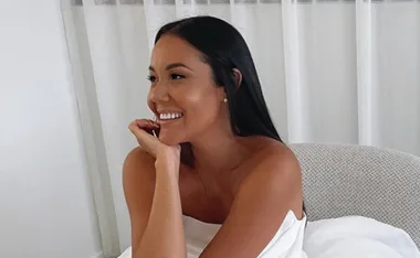 A woman with long dark hair smiling and seated, wearing a strapless dress indoors.