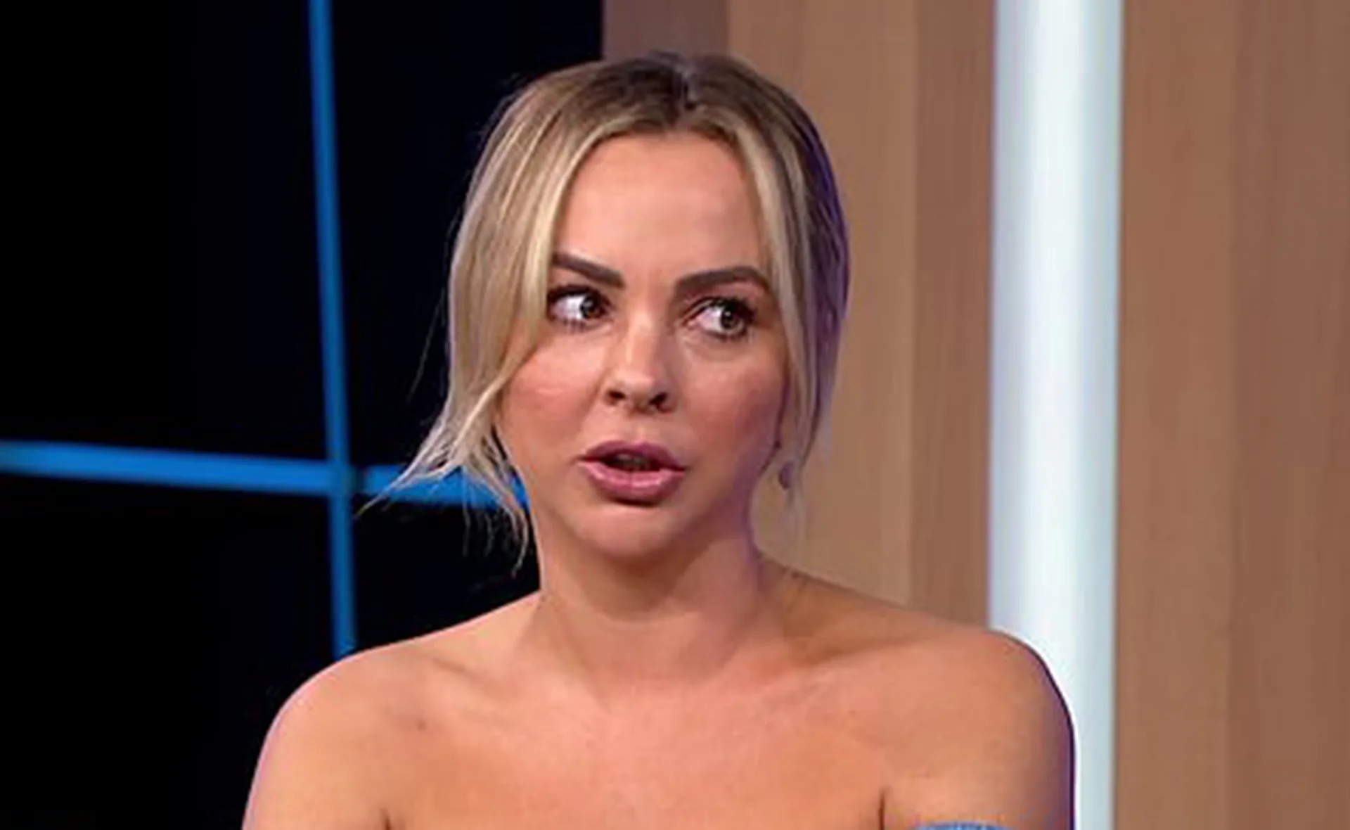 A woman with blonde hair appears to be speaking during a TV interview, wearing an off-shoulder outfit.