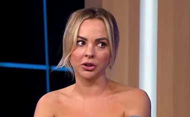 A woman with blonde hair appears to be speaking during a TV interview, wearing an off-shoulder outfit.