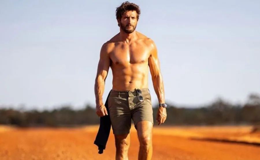Shirtless man walking on a red dirt road, holding a jacket, wearing shorts and sunglasses. Background of clear sky and trees.