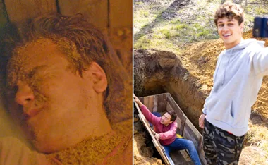 A man covered in sand is buried alive; another man takes a selfie near an open wooden box in the ground.