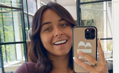 A person with shoulder-length brown hair taking a selfie in a bright room, smiling and holding a phone with a monogrammed case.