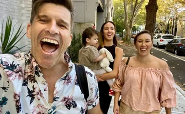 Man in floral shirt and three people, including a woman holding a child with a stuffed toy, smiling outdoors on a tree-lined street.