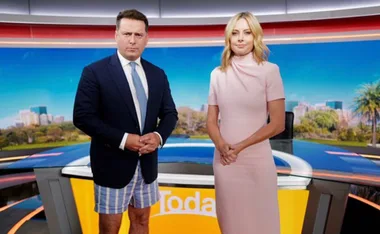 TV hosts on set; one in a suit jacket and shorts, the other in a pink dress, standing in front of a backdrop.