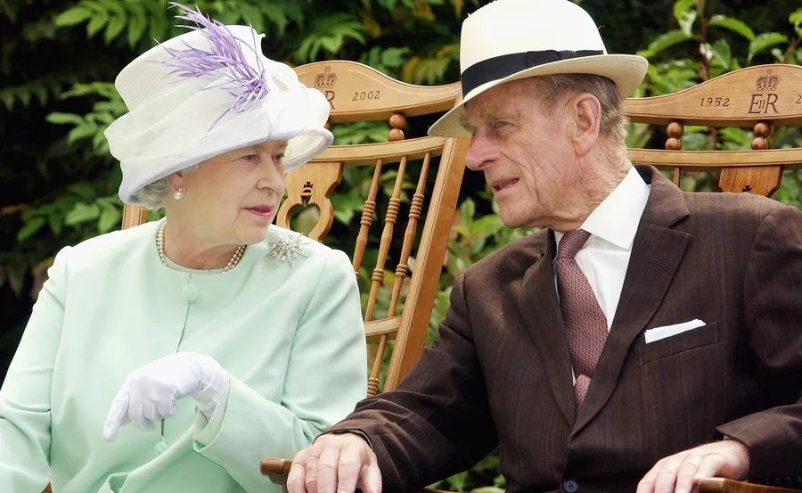 Two elderly people wearing hats, sitting outdoors on wooden chairs, engaged in conversation.