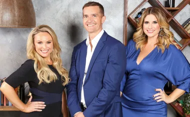 Group of three people smiling, two women and one man, dressed in formal attire, against a bookshelf background.