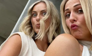 Two women with blonde hair make playful duck faces in a selfie indoors.