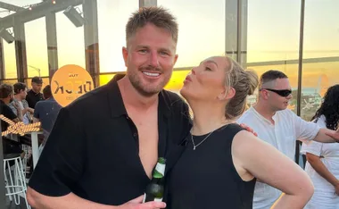 Man smiling with drink and woman in black dress at sunset, rooftop bar setting.