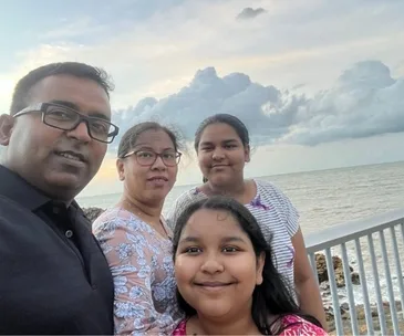 A family of four smiling on a seaside boardwalk, with the ocean and a cloudy sky in the background.