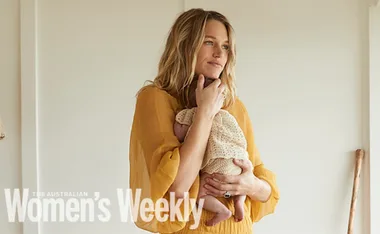 A woman in a yellow dress holds a baby wrapped in a cream blanket, with "Women's Weekly" text on the image.