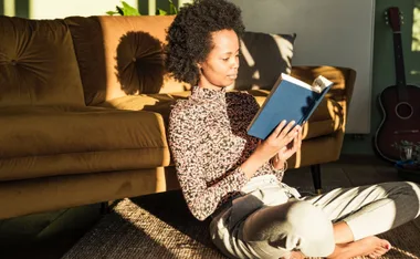 Woman sitting on floor reading a book in sunlight, with a guitar nearby.