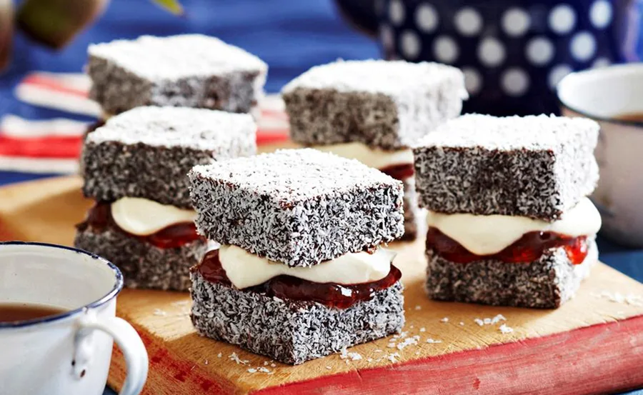 Lamingtons filled with cream and jam on a wooden board, with cups of tea in the background.