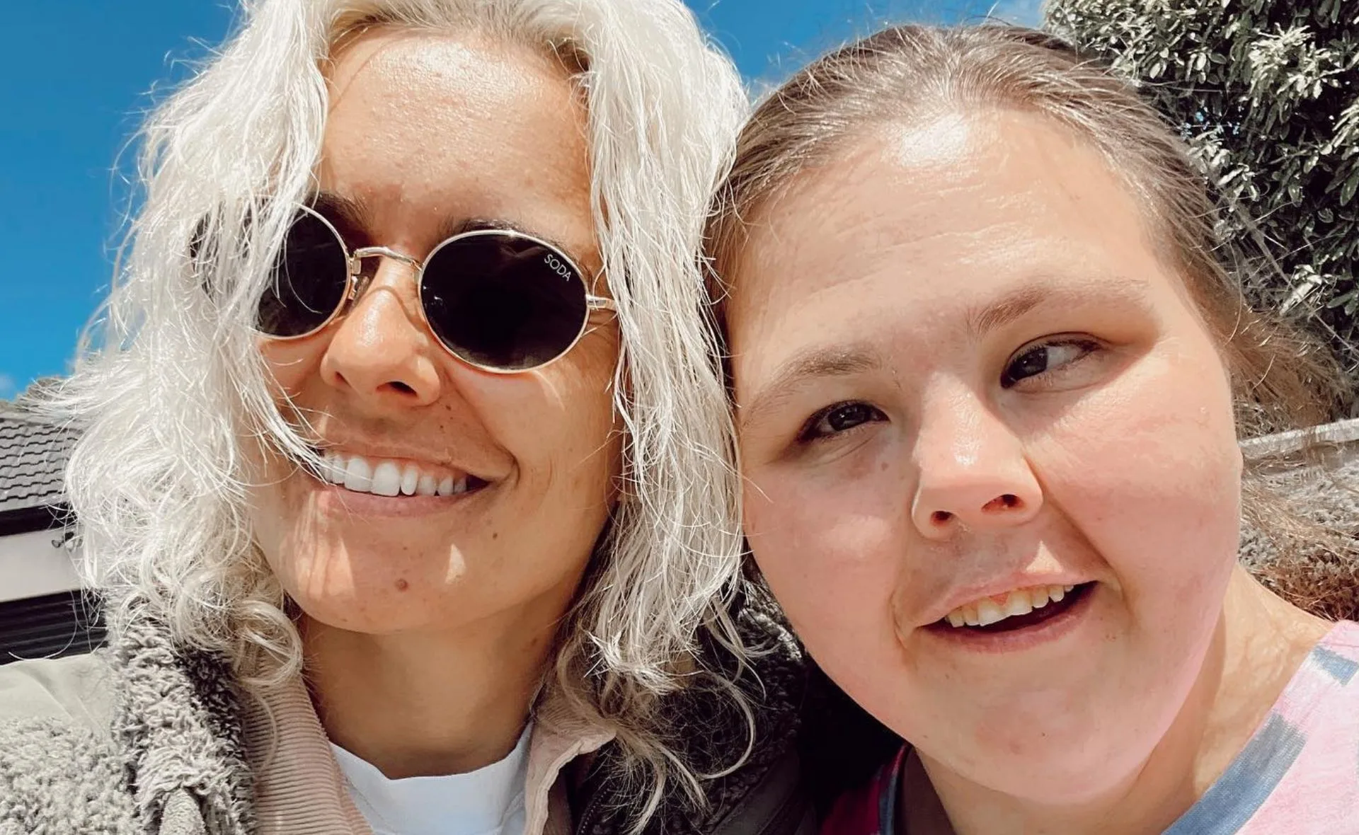Two people smiling for a selfie outdoors, one with sunglasses and white hair, under a clear blue sky.