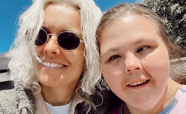 Two people smiling for a selfie outdoors, one with sunglasses and white hair, under a clear blue sky.