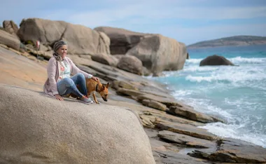 Woman sitting on rocks by the ocean with a dog, rocky coastline and waves in the background.