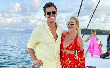 A man in a yellow shirt and a woman in a red dress smiling on a boat, with people and the ocean in the background.