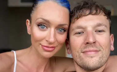 Two people smiling closely together in a selfie, wearing casual attire indoors.