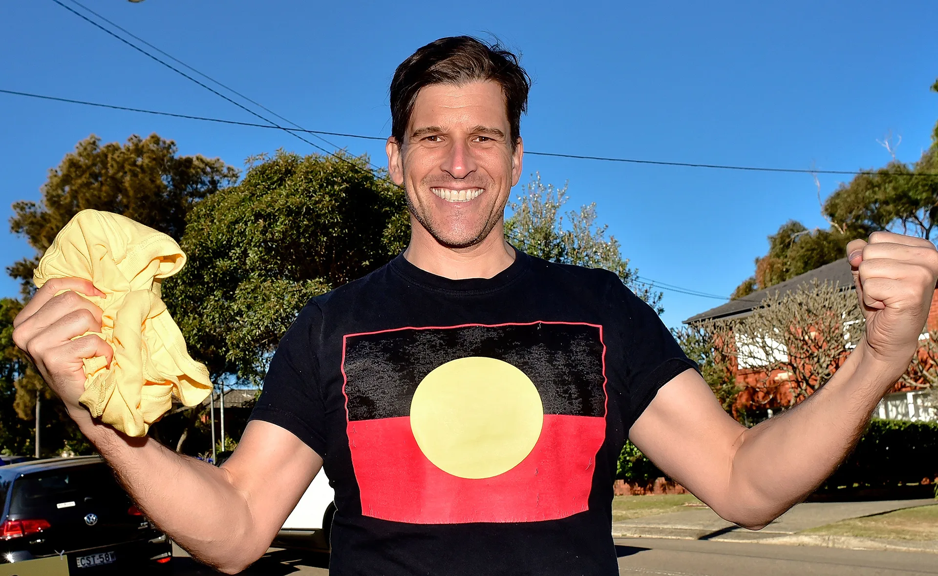 Man smiling and wearing a black shirt with an Aboriginal flag, holding a yellow cloth, outdoors with trees in background.