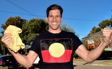 Man smiling and wearing a black shirt with an Aboriginal flag, holding a yellow cloth, outdoors with trees in background.