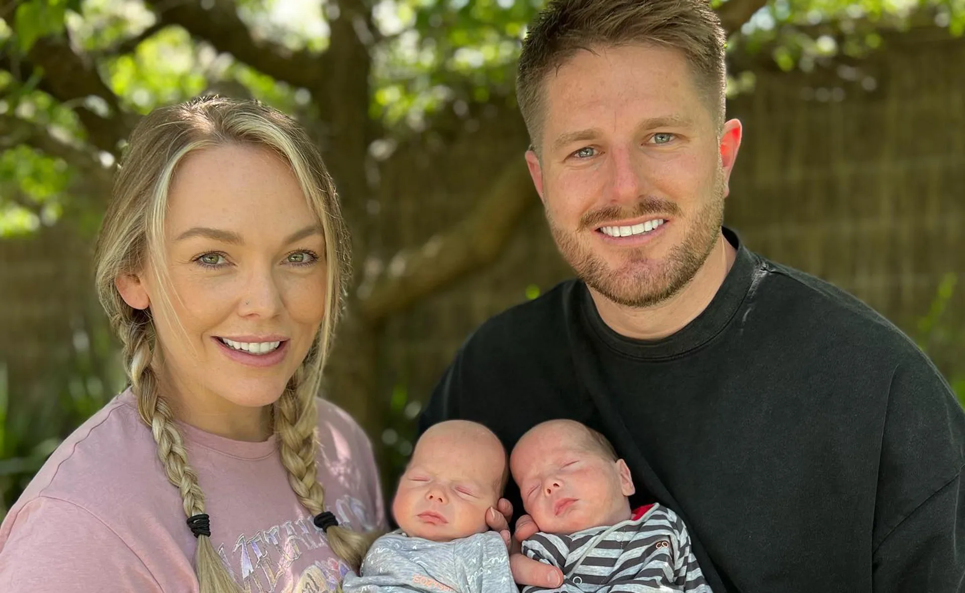 A couple smiling outdoors, each holding a sleeping baby under a tree.
