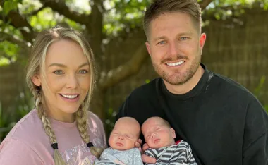 A couple smiling outdoors, each holding a sleeping baby under a tree.