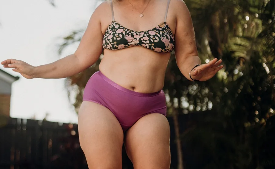 Person wearing a floral bikini top and purple high-waisted period underwear, outside near greenery.