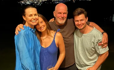 A group of four smiling people standing close together at night with city lights in the background.