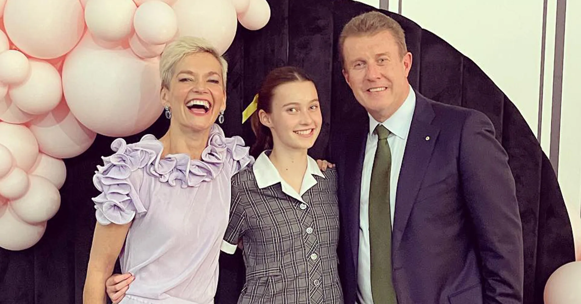 A joyful family poses together; a woman in a lilac dress, a young girl in a school uniform, and a man in a suit with pink balloons.