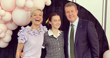 A joyful family poses together; a woman in a lilac dress, a young girl in a school uniform, and a man in a suit with pink balloons.