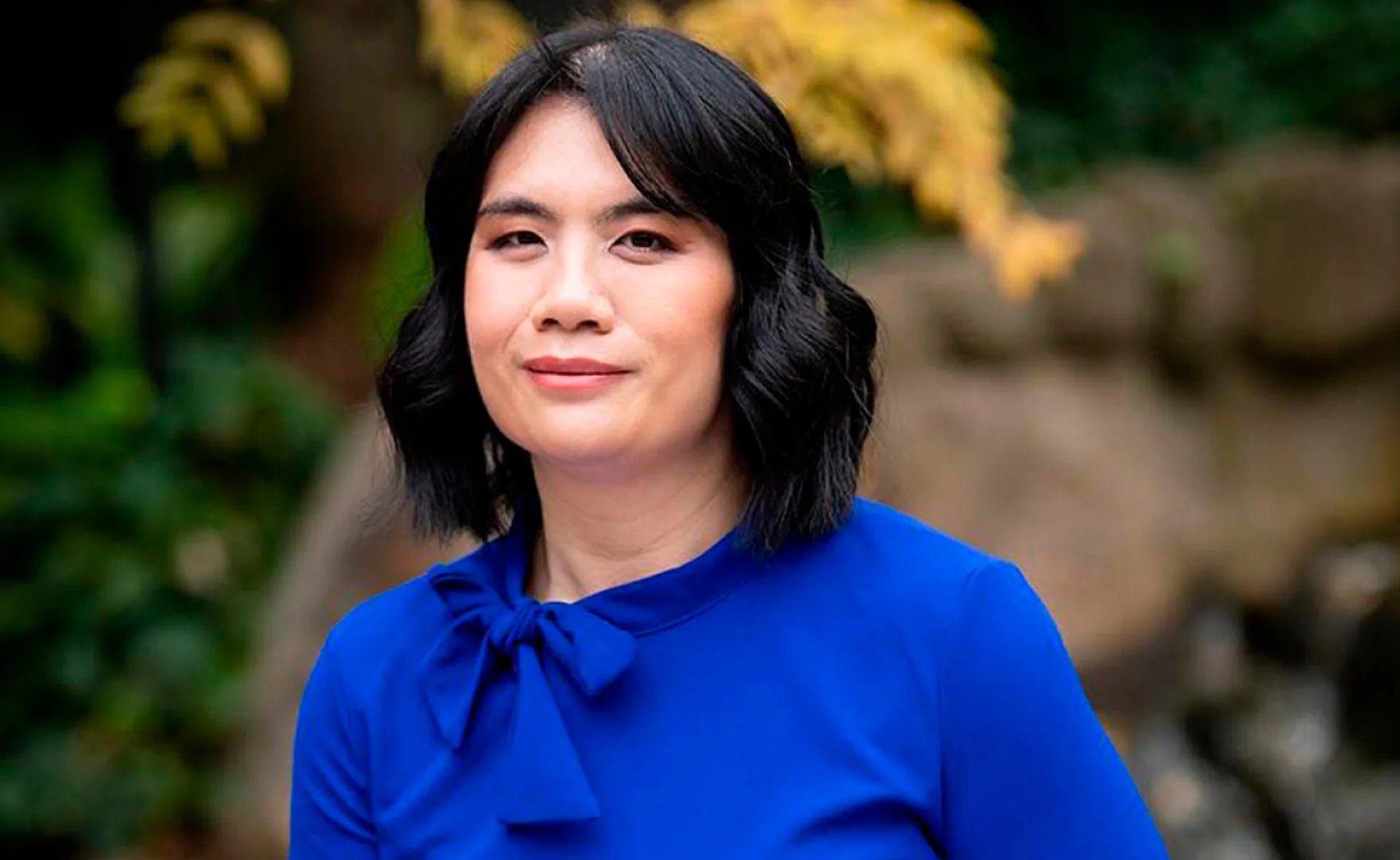 A woman with dark hair in a blue outfit stands outdoors with blurred greenery in the background.