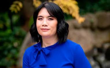 A woman with dark hair in a blue outfit stands outdoors with blurred greenery in the background.