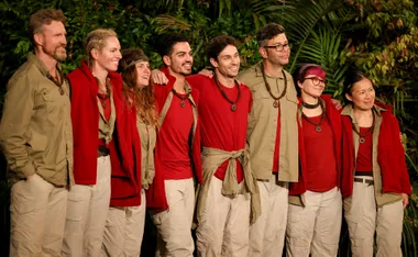 Contestants in red and beige outfits from "I'm a Celebrity 2022" standing together, smiling in the jungle.