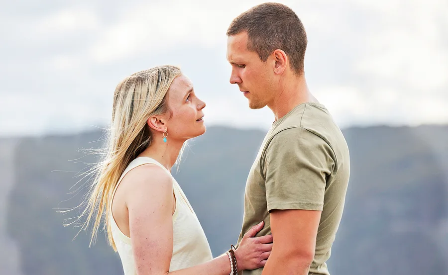 A couple stands closely facing each other outdoors, the woman touches the man's chest, both looking serious.