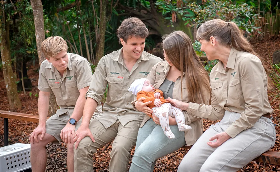 A family sits on a bench outdoors, wearing khaki zoo uniforms, holding a baby dressed in orange and white.
