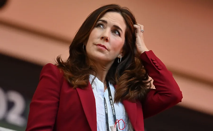A woman with long brown hair in a red blazer looks pensive as she touches her hair.