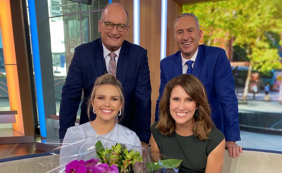 Four smiling people in a TV studio; two standing behind a couch and two sitting in front holding a bouquet of flowers.