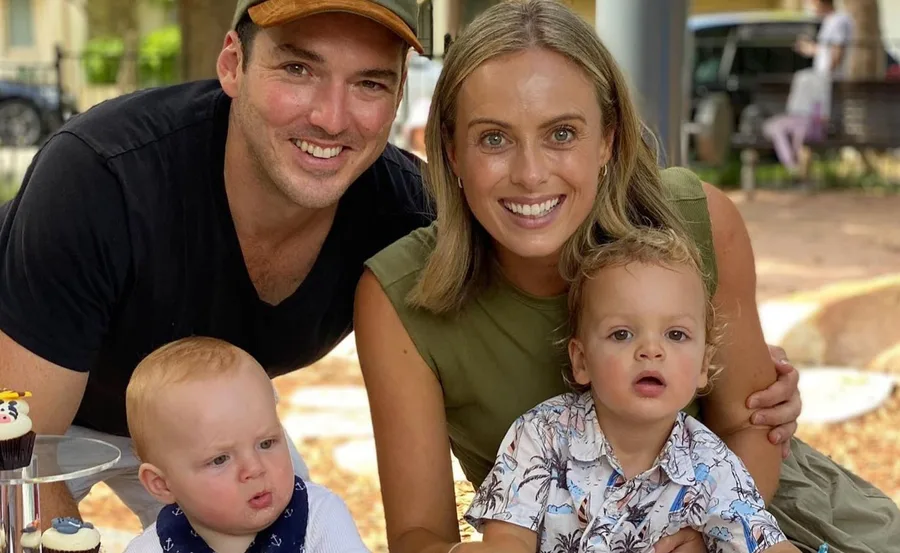 A family of four smiles outdoors, with two young children, one in floral shirt, by a stand of cupcakes.