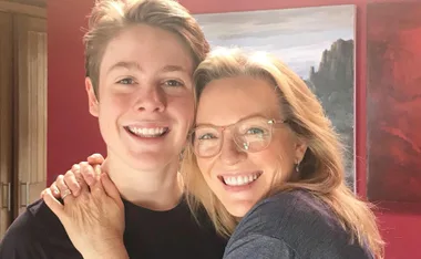 A woman with glasses and a young man smiling, standing close together indoors with paintings in the background.