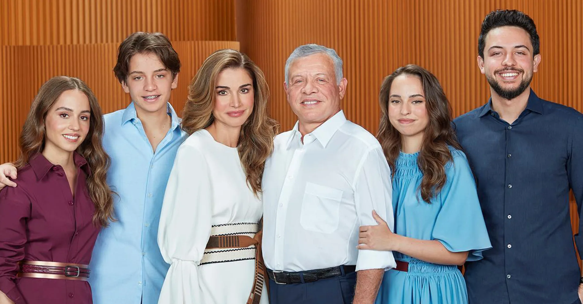 A family portrait with six people standing close, smiling, in formal attire, with a wooden panel background.