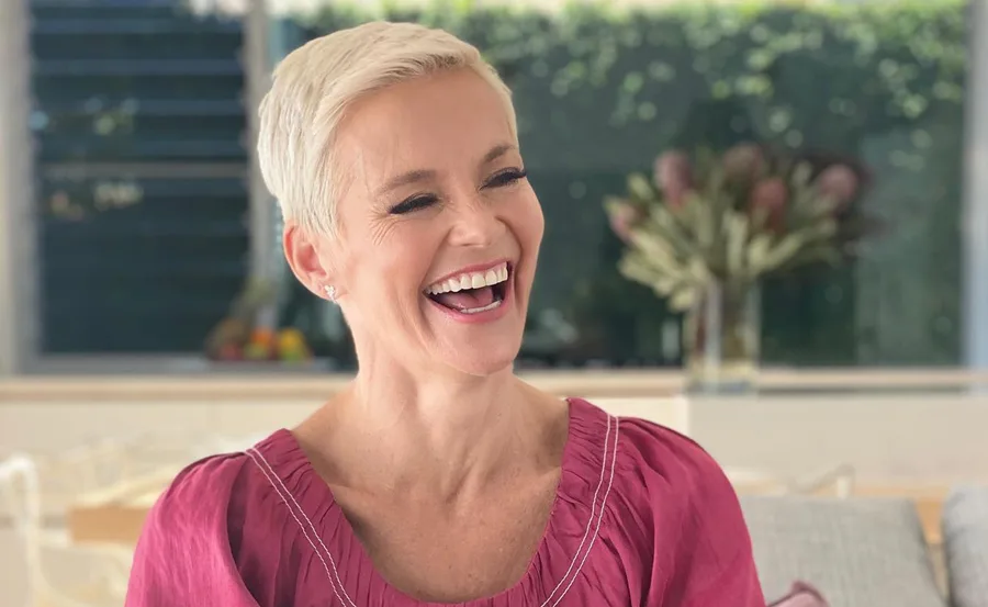 A person with short blonde hair, wearing a pink top, is laughing in a warmly lit room.