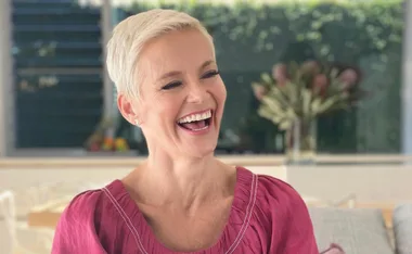 A person with short blonde hair, wearing a pink top, is laughing in a warmly lit room.