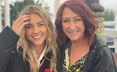 Two women smiling outdoors, one with blonde hair, the other with red hair, both wearing casual jackets.