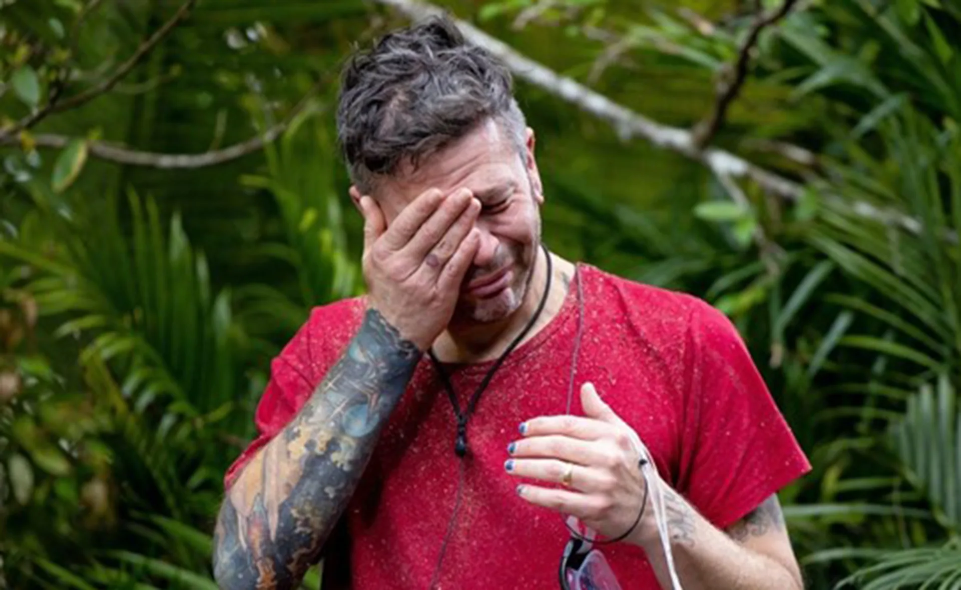 Person in a red shirt with tattoos, looking emotional and wiping tears in a jungle setting.
