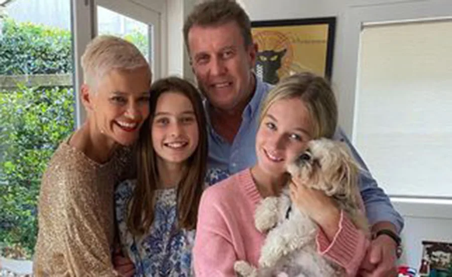A family smiling together, with two children and a dog, standing inside near a window.