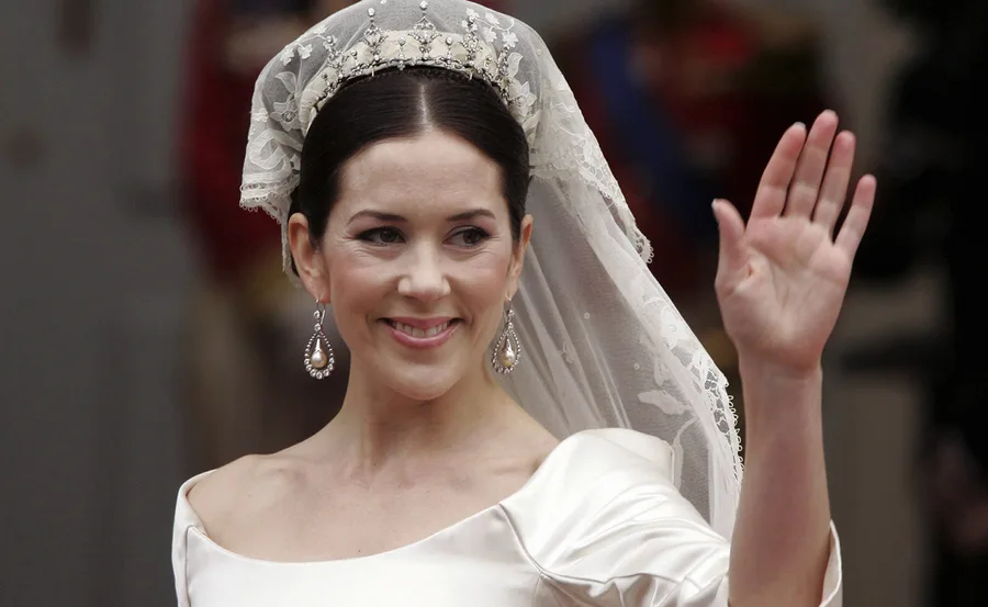 A bride in a white wedding dress and veil smiles and waves, wearing a tiara and pearl earrings.