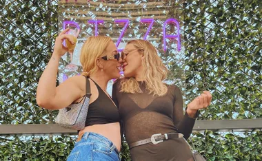 Two women in stylish outfits nearly kissing, with a backdrop of greenery and "PIZZA" in neon lights behind them.