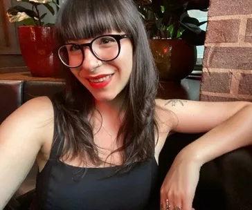 A woman with glasses and long hair smiles indoors, sitting by a brick wall and plants.