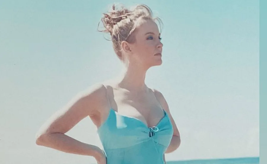 A woman in a blue dress stands with hands on hips by the sea, under a clear sky.