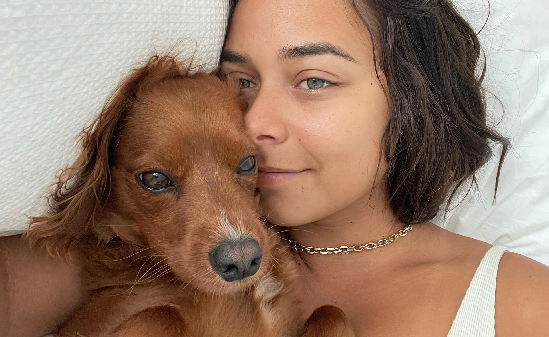 A woman lying on white sheets with a small brown dog, both looking at the camera.