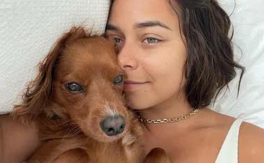 A woman lying on white sheets with a small brown dog, both looking at the camera.